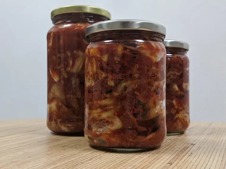 Three glass jars filled with red-seasoned cabbage kimchi on a wooden surface.