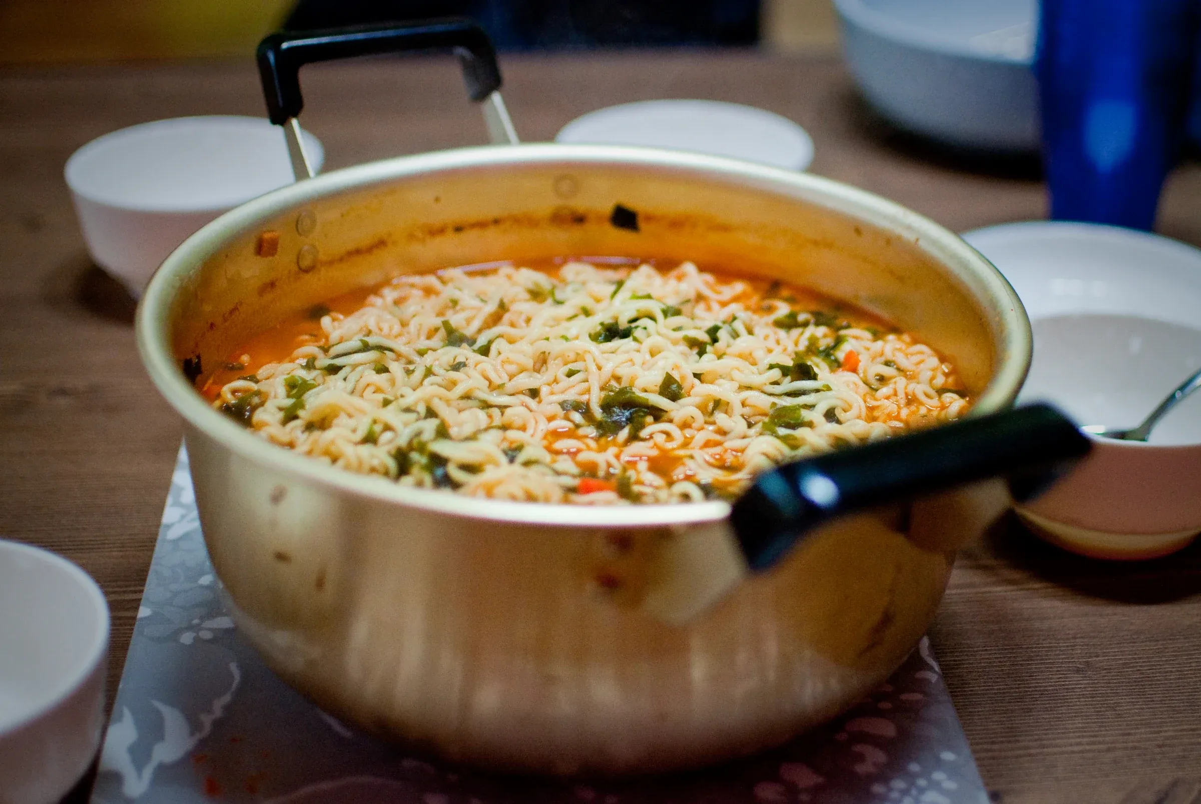 Cooked instant ramyeon noodles in reddish broth with green vegetables, served in a gold-colored metal pot.