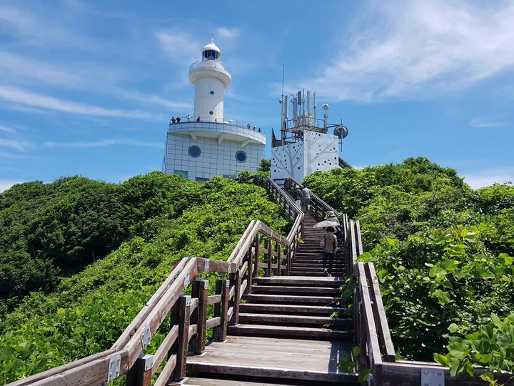 A wooden stairway climbs through green vegetation to the white lighthouse on Ongdo, Taean.