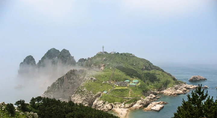 Mist drifts around rocky cliffs beside a green lighthouse islet viewed from Somaemuldo.